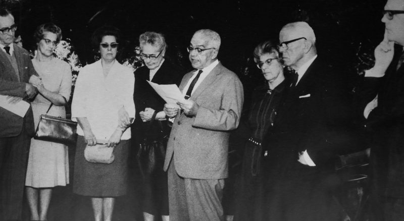 Foto preto e branca de 1964 mostra um conjunto de oito pessoas, entre homens e mulheres adultos. Ao centro, um homem grisalho e de terno, lê com um papel na mão enquanto os outros o observam